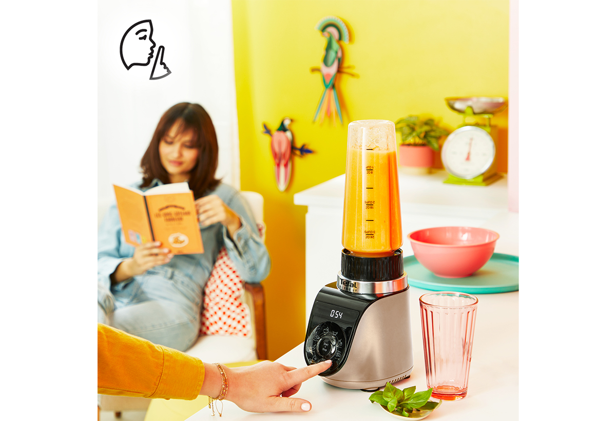 Απεικονίζεται μια γυναίκα να χρησιμοποιεί το μπλέντερ, ενώ μια άλλη γυναίκα διαβάζει ένα βιβλίο στο ίδιο δωμάτιο.